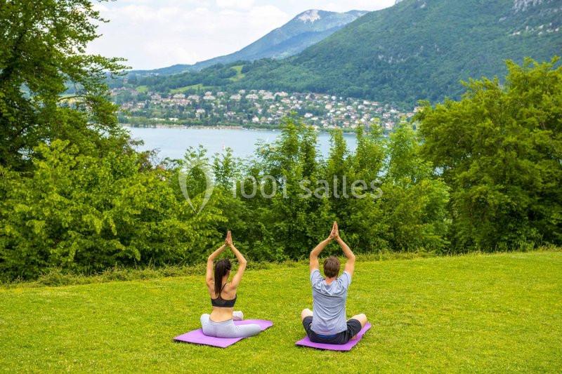 Deux personnes pratiquent le yoga sur des tapis dans un parc verdoyant avec un lac et des montagnes en arrière-plan.