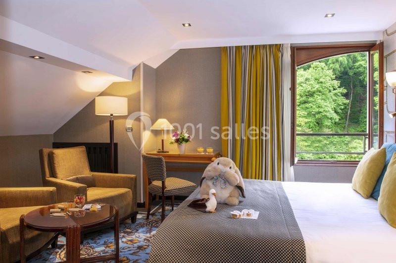 Chambre lumineuse avec lit, fauteuils, table basse, fenêtre ouverte sur un paysage verdoyant et décoration soignée.