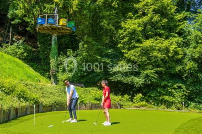 Deux personnes jouent au mini-golf sur un terrain entouré de végétation, avec une cabane perchée visible en arrière-plan.