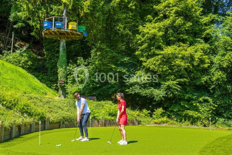 Deux personnes jouent au mini-golf sur un terrain entouré de végétation, avec une cabane perchée visible en arrière-plan.