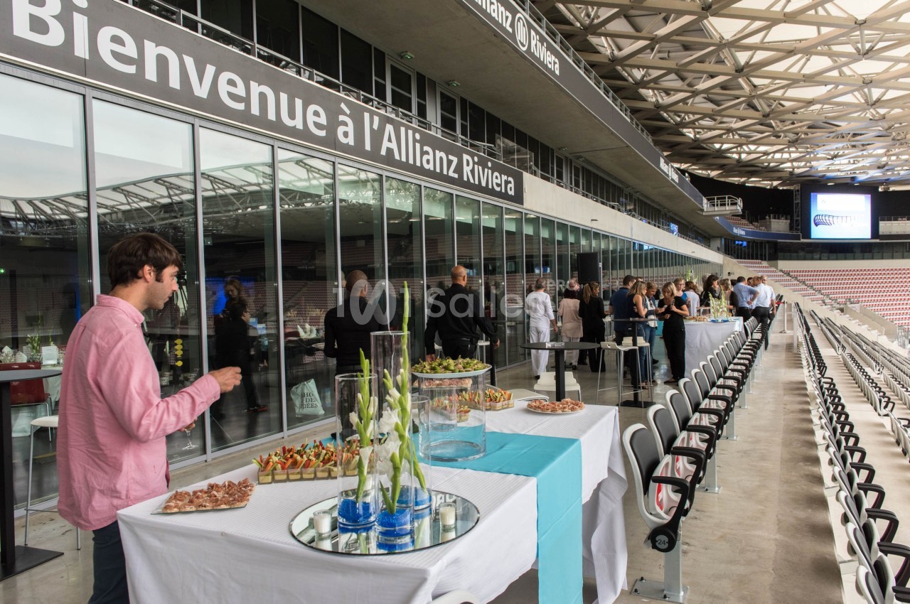 Des invités se tiennent près d'une table de buffet dans un espace événementiel avec vue sur des gradins de stade.