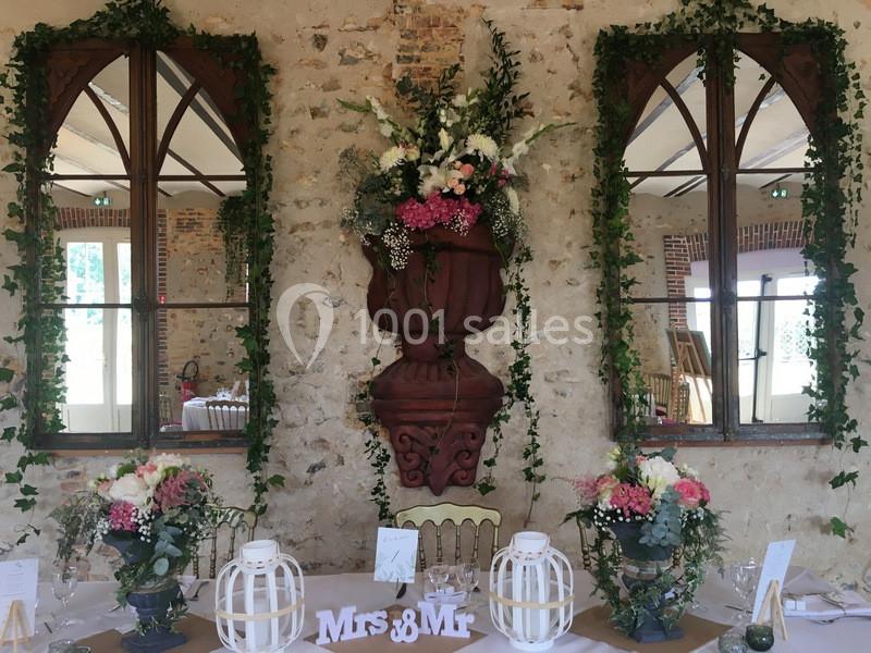 Décoration de mariage avec fleurs, feuillages et lanternes blanches sur une table devant un mur en pierre et deux miroirs.