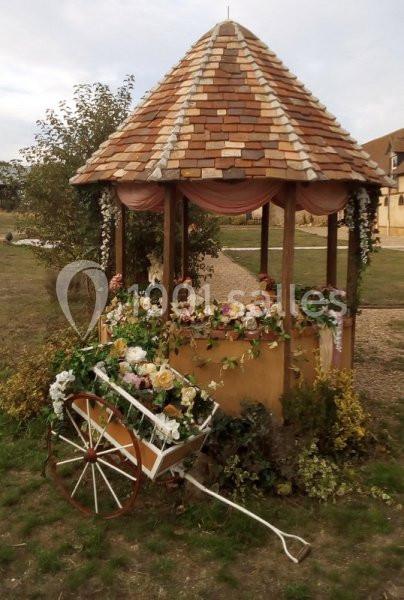 Kiosque en bois avec toit en tuiles, décoré de fleurs et accompagné d'une charrette fleurie sur une pelouse.