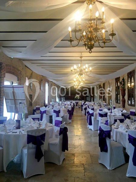 Salle de réception décorée pour un événement, avec des tables dressées et des chaises ornées de nœuds violets.
