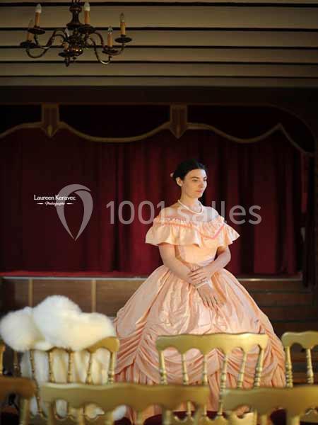 Femme en robe rose à volants debout sur une scène avec rideaux rouges, dans une salle ornée d'un lustre.