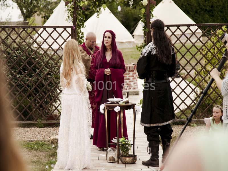 Cérémonie en plein air avec une femme en robe rouge officiant devant deux personnes en tenue médiévale.