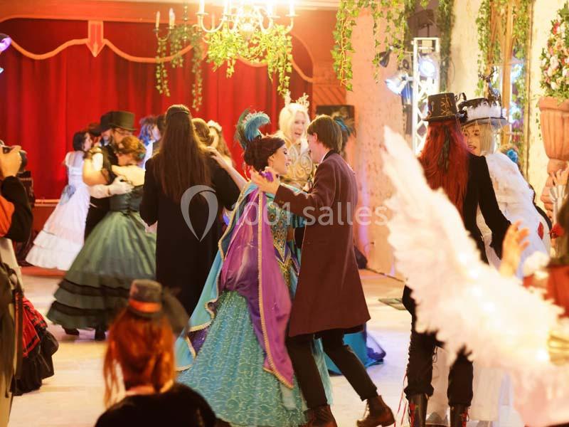 Des personnes en costumes d'époque dansent dans une salle ornée de rideaux rouges et de décorations végétales.