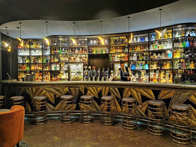 Bar élégant avec comptoir en bois sculpté, tabourets alignés et étagères remplies de bouteilles illuminées.