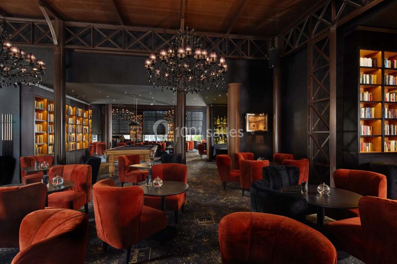 Salon élégant avec fauteuils rouges, tables rondes, étagères remplies de livres et lustres noirs suspendus.