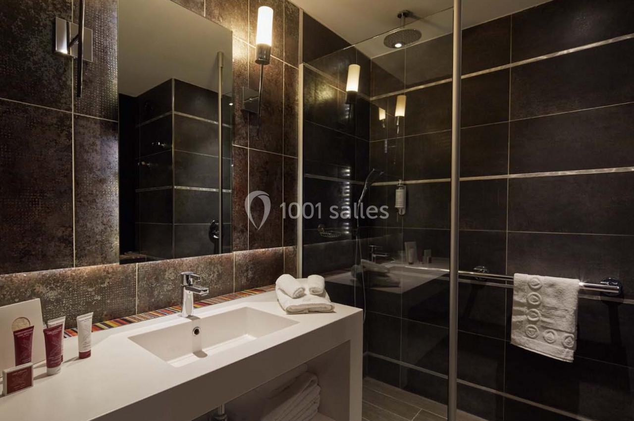 Salle de bain moderne avec lavabo blanc, douche vitrée, murs carrelés sombres et éclairage chaleureux.
