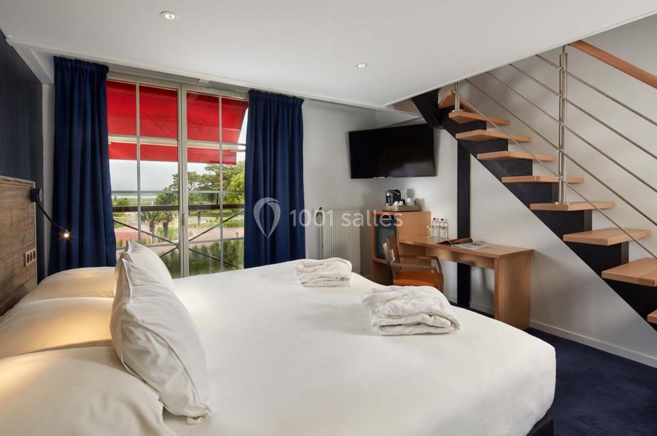 Chambre lumineuse avec lit double, bureau, télévision, escalier en bois et grande fenêtre avec rideaux bleus.
