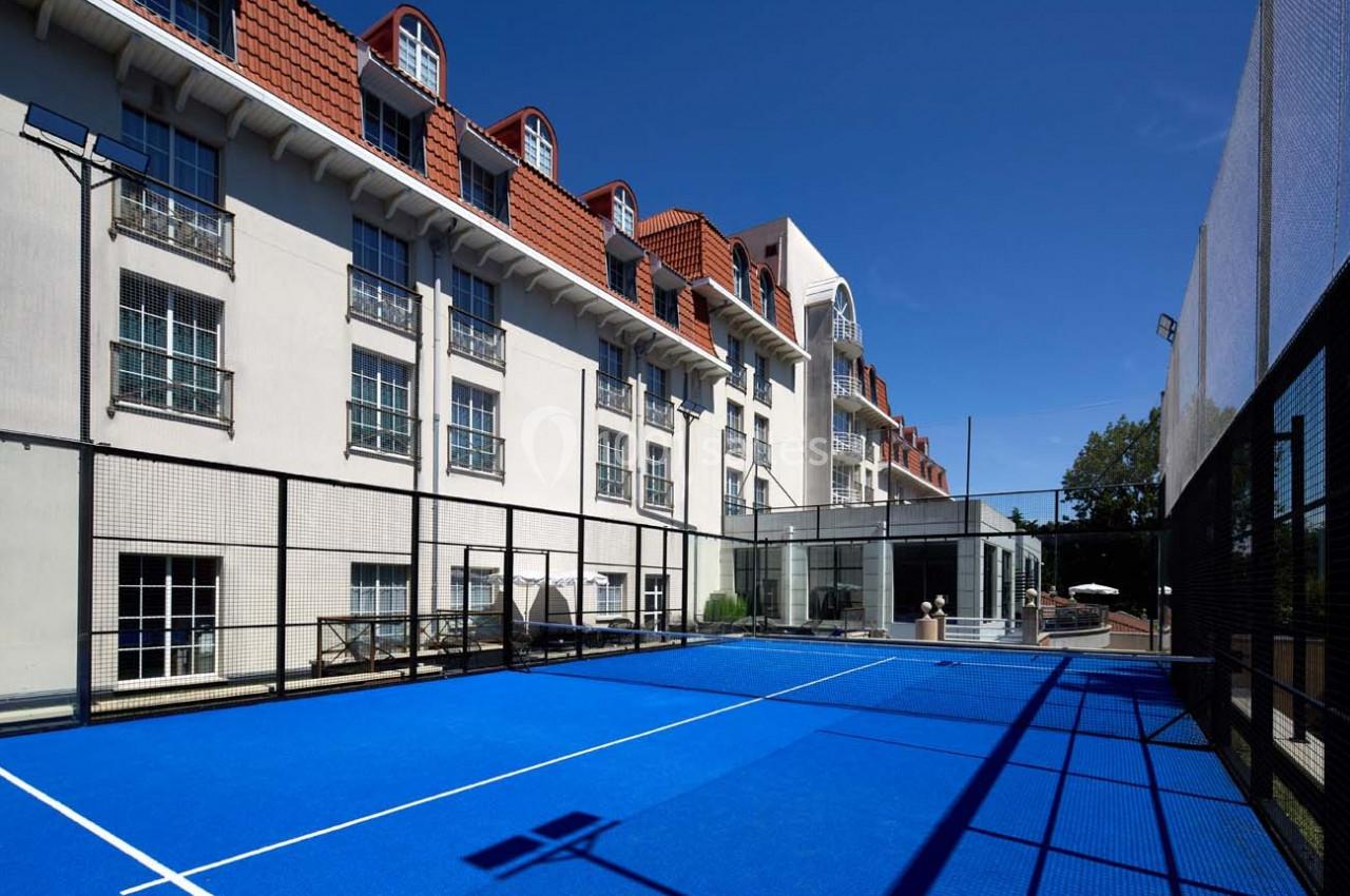 Terrain de padel extérieur avec sol bleu, entouré de filets, situé devant un bâtiment à plusieurs étages.