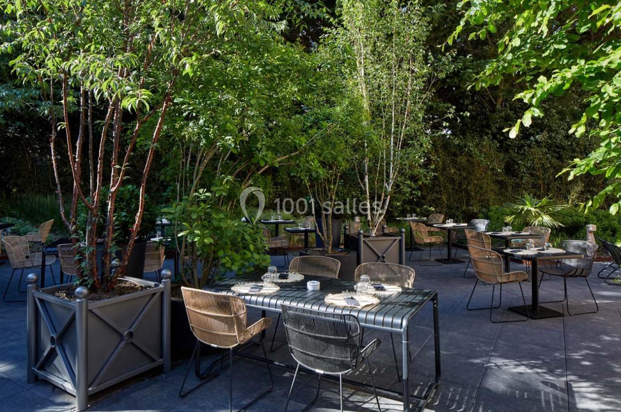 Terrasse extérieure avec tables et chaises entourées de plantes et d'arbres, dans un cadre verdoyant.