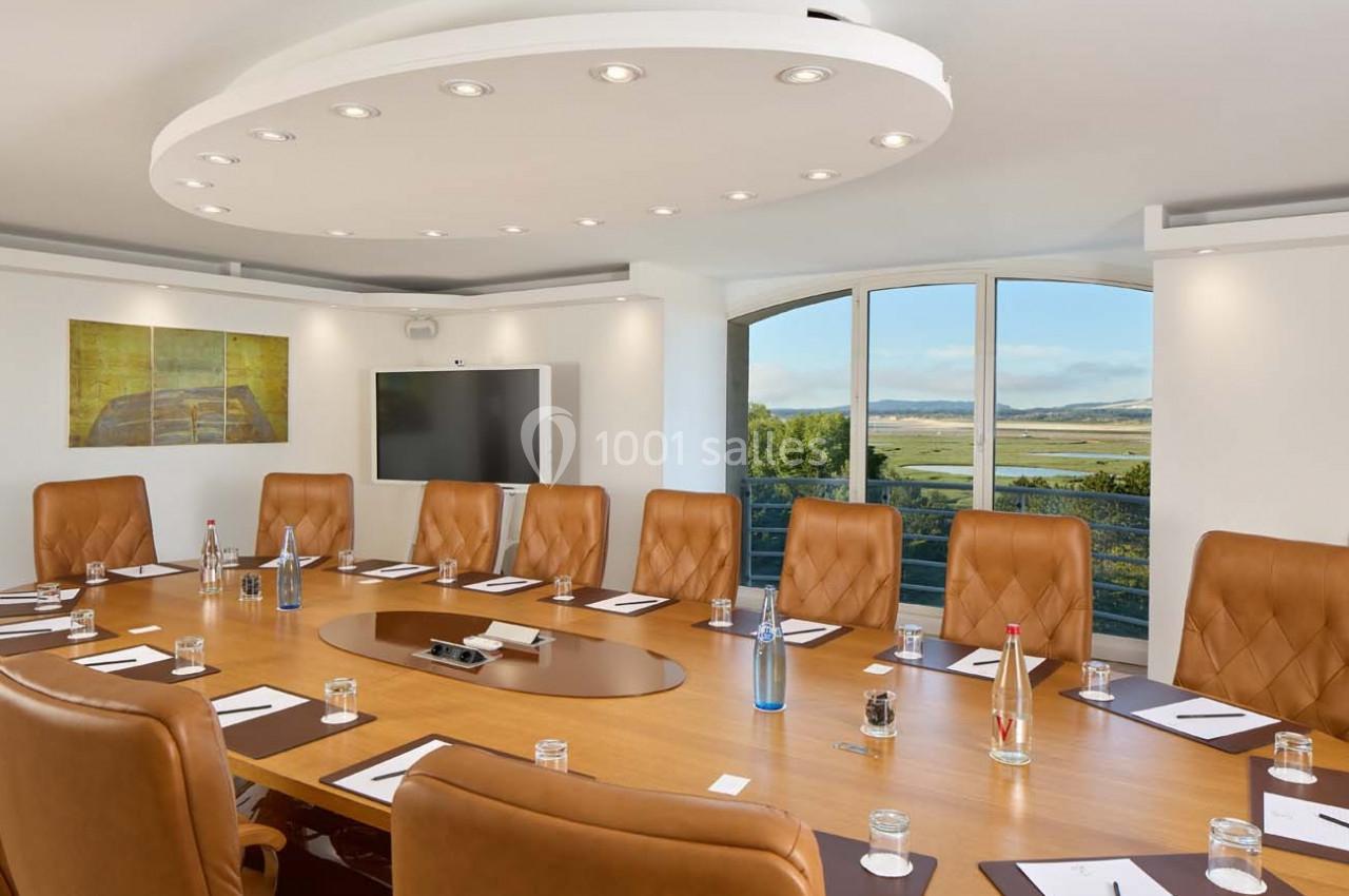 Salle de réunion lumineuse avec table ovale en bois, chaises en cuir, écran mural et vue sur un paysage extérieur.