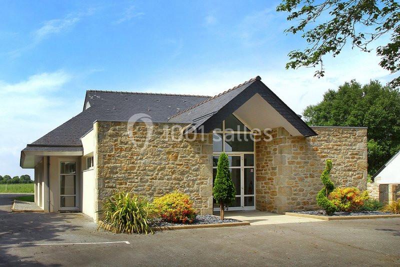 Bâtiment moderne en pierre et ardoise entouré de végétation, avec une entrée vitrée et un ciel dégagé.