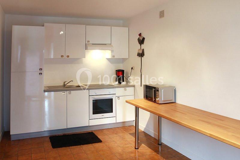 Cuisine équipée avec meubles blancs, évier, four, hotte, micro-ondes sur une table en bois et carrelage au sol.