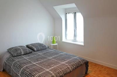 Chambre lumineuse avec un lit double couvert d'une housse à motifs noirs et blancs, près d'une fenêtre avec rideaux clairs.