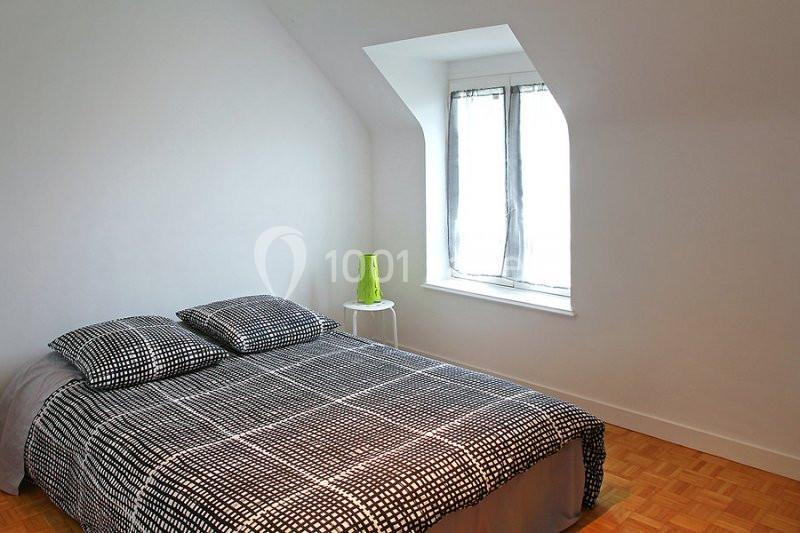 Chambre lumineuse avec un lit double couvert d'une housse à motifs noirs et blancs, près d'une fenêtre avec rideaux clairs.