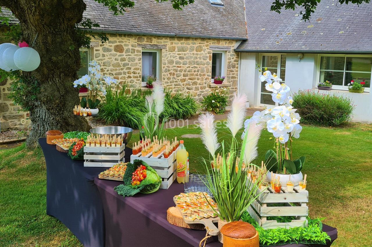 Table de buffet en extérieur avec plats variés, fleurs décoratives et lanternes suspendues, devant une maison en pierre.