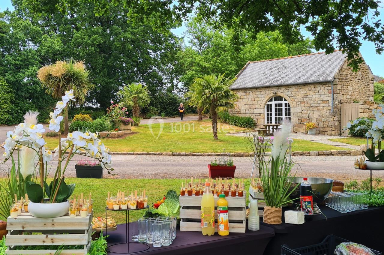 Table de buffet en extérieur avec boissons et fleurs, devant un jardin verdoyant et une bâtisse en pierre.
