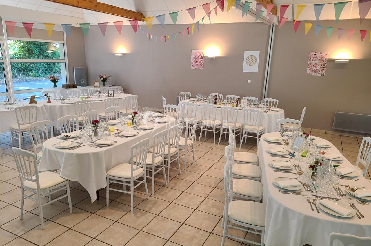 Salle décorée pour un événement avec des tables dressées, des chaises blanches et des guirlandes colorées suspendues.
