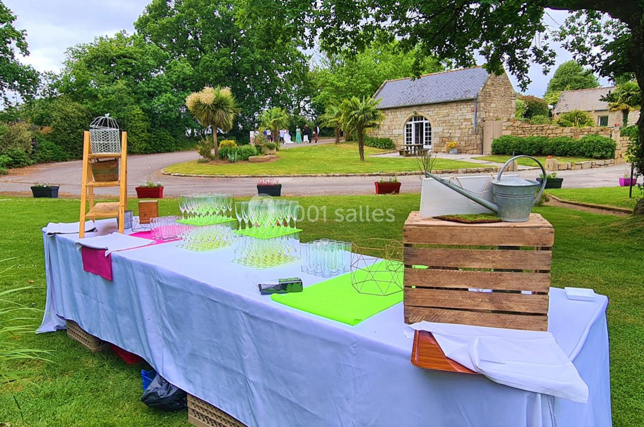 Table dressée en extérieur avec verres, décorations et arrosoir, dans un jardin près de bâtiments en pierre.