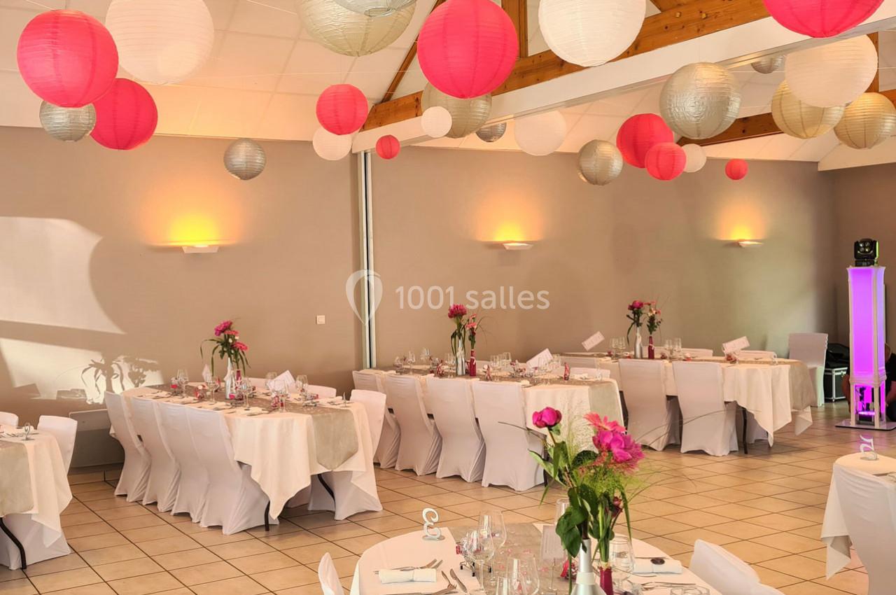 Salle de réception décorée avec des lanternes suspendues et des tables dressées avec nappes blanches et fleurs roses.
