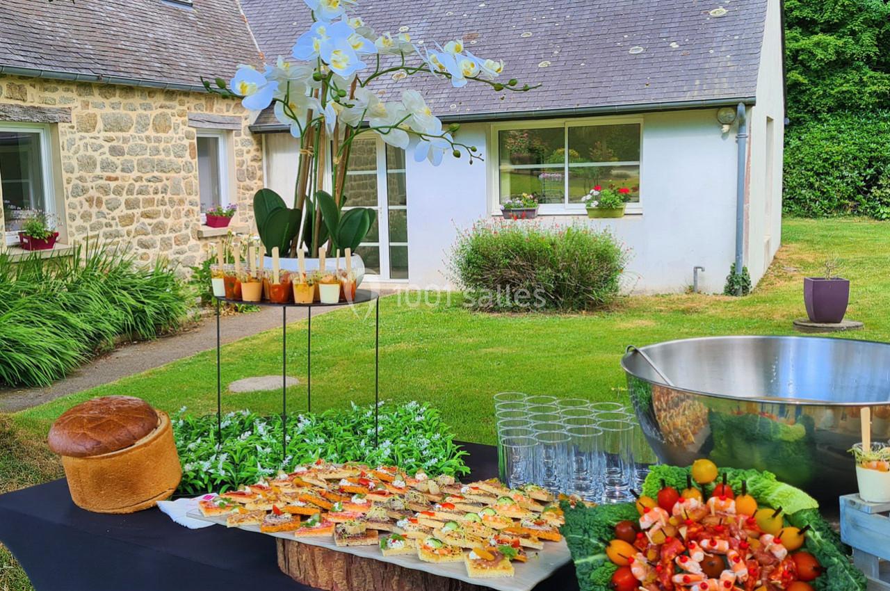 Table dressée en extérieur avec des amuse-bouches, un pain, des verrines et des fleurs, devant une maison en pierre.