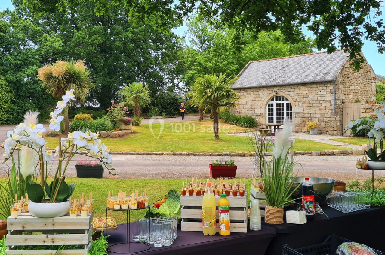 Buffet en extérieur avec boissons et amuse-bouches, devant un jardin verdoyant et une petite maison en pierre.