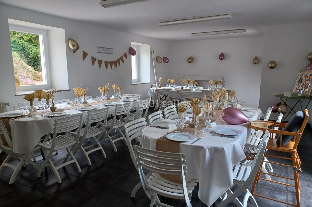Salle décorée pour un événement avec tables dressées, nappes blanches, chaises pliantes et décorations dorées et roses.