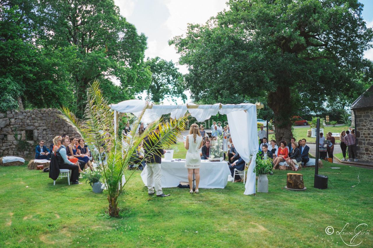 Cérémonie en plein air sous une arche décorée de voilages blancs, entourée d'invités assis sur une pelouse verdoyante.