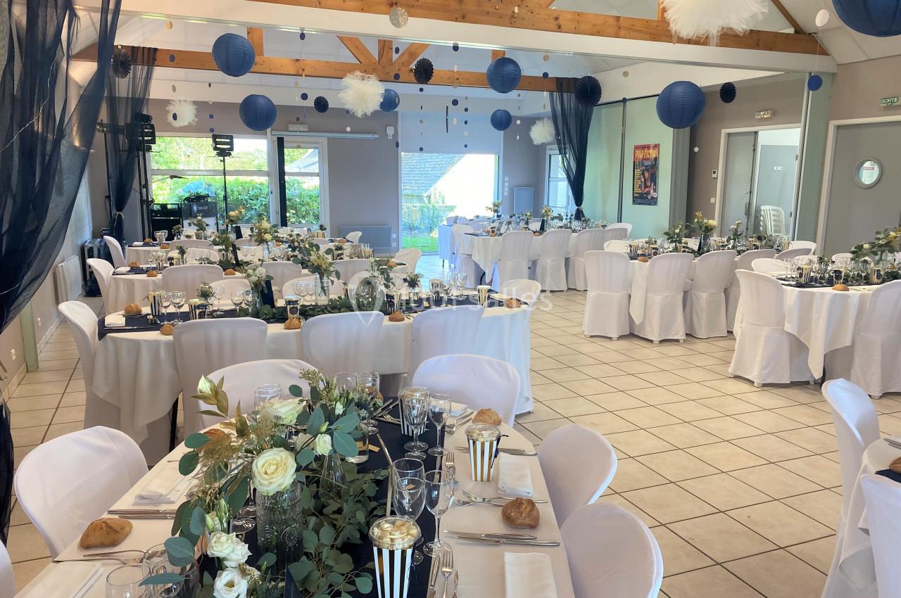 Salle de réception décorée pour un événement, avec tables dressées, fleurs, nappes blanches et lanternes suspendues.