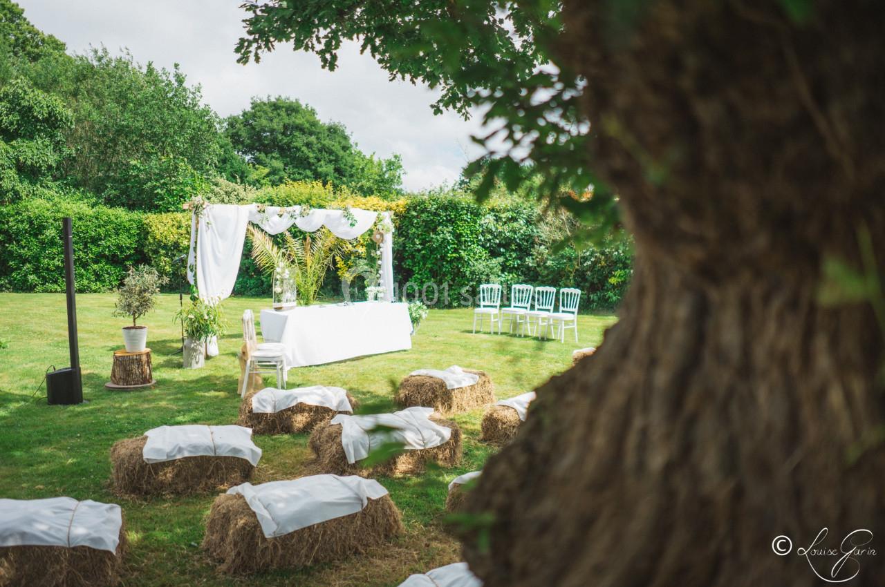 Cérémonie en plein air avec une arche décorée de fleurs, des bottes de paille et des chaises disposées sur une pelouse.