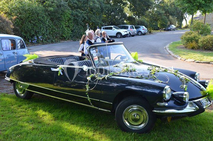 Voiture ancienne décapotable noire décorée de fleurs, stationnée sur une pelouse près d'une route bordée d'arbres.