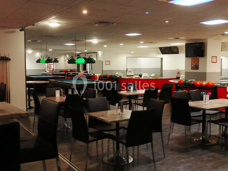 Salle de bowling avec tables, chaises noires, billards éclairés et rangée de boules colorées en arrière-plan.