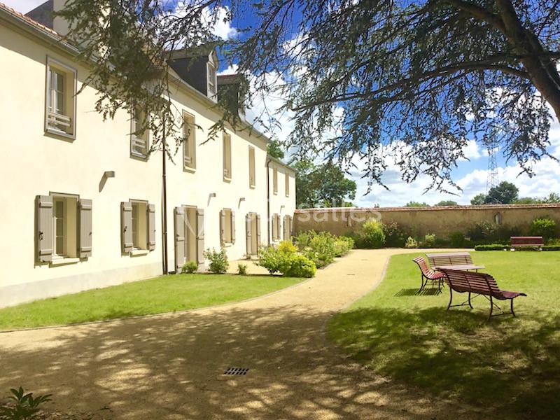 Façade d'un bâtiment clair entouré d'un jardin avec pelouse, bancs et allée sous un ciel bleu.