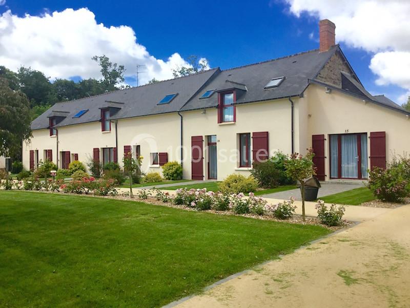 Grande maison de campagne avec façade claire, volets rouges, jardin fleuri et allées gravillonnées sous un ciel bleu.