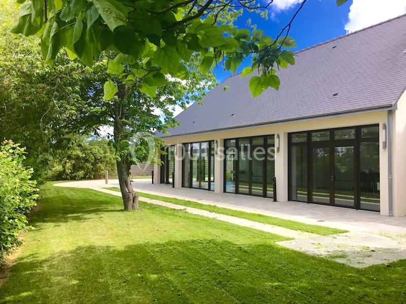 Maison moderne avec de grandes baies vitrées donnant sur un jardin verdoyant et un ciel bleu dégagé.