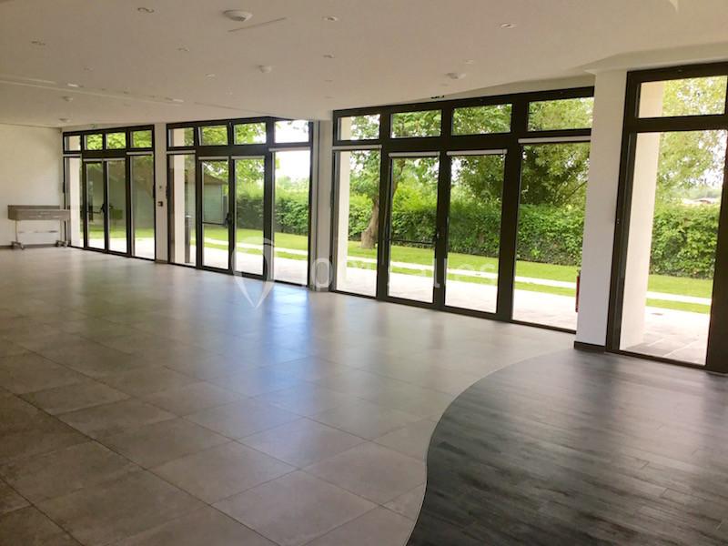 Salle lumineuse avec de grandes baies vitrées donnant sur un jardin verdoyant, sol en carrelage et parquet.