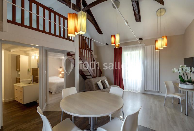 Intérieur lumineux d'un appartement avec mezzanine, table ronde, canapé, chambre visible et poutres apparentes au plafond.