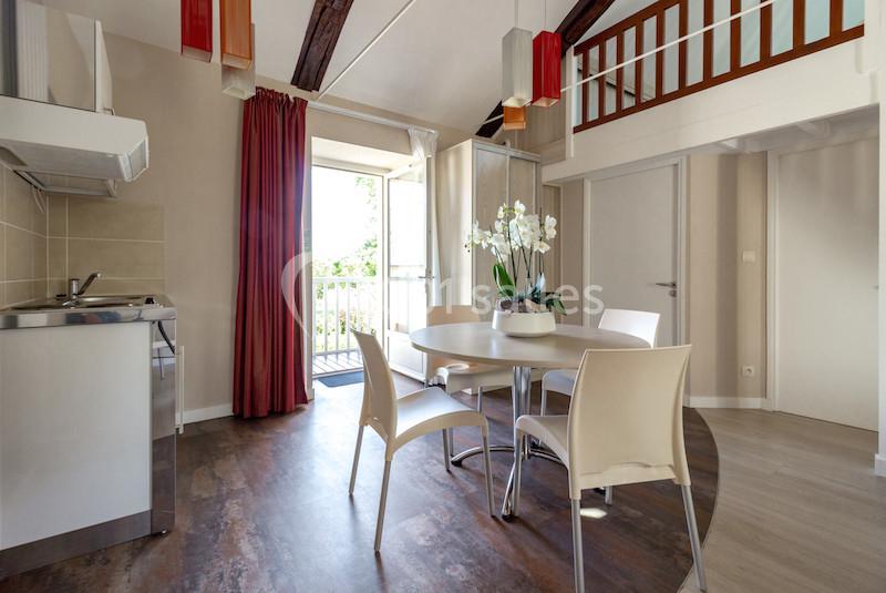 Salle à manger lumineuse avec table ronde, chaises blanches, cuisine ouverte et balcon donnant sur l'extérieur.