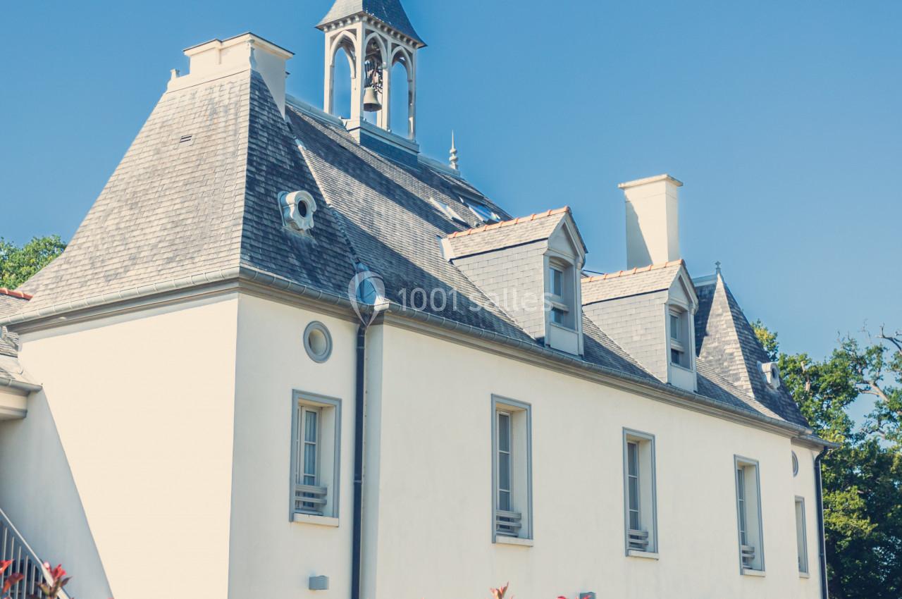 Bâtiment blanc à toit en ardoise avec un petit clocher, entouré de verdure sous un ciel bleu.