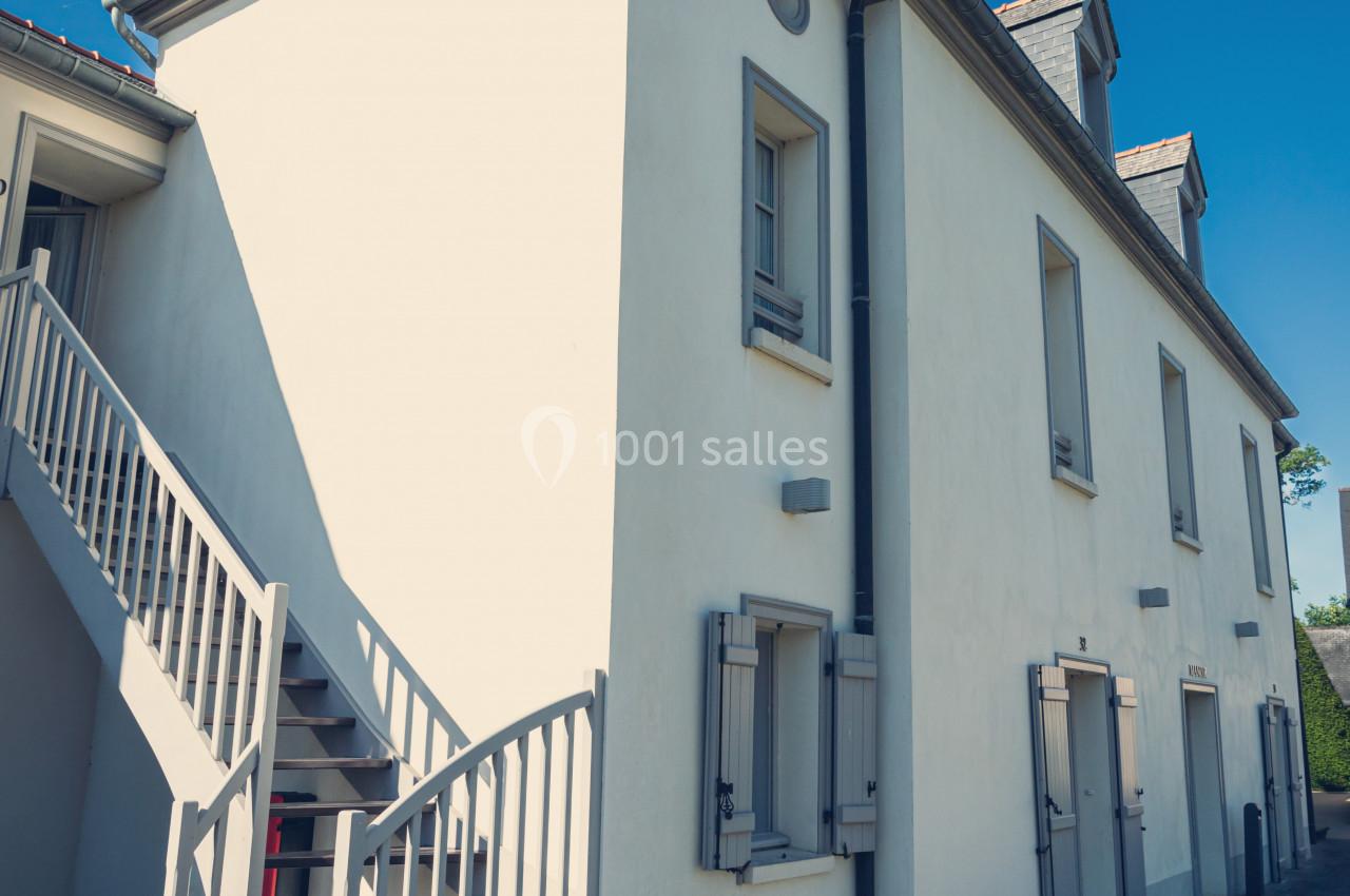 Façade d'une maison blanche à deux étages avec escalier extérieur en bois et toit en ardoise sous un ciel bleu.