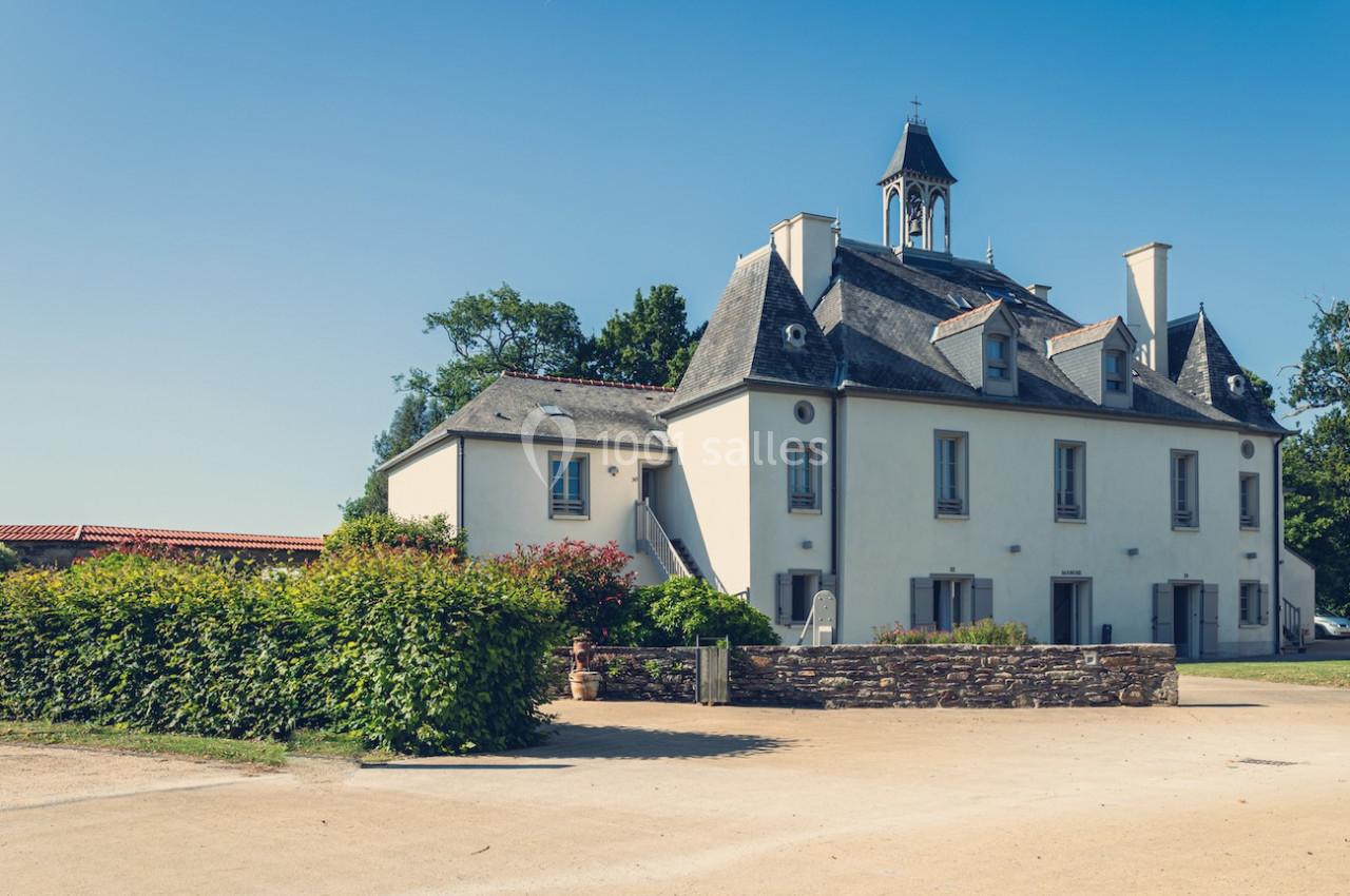 Bâtiment ancien à deux étages avec toit en ardoise, entouré de verdure et baigné de lumière sous un ciel bleu.