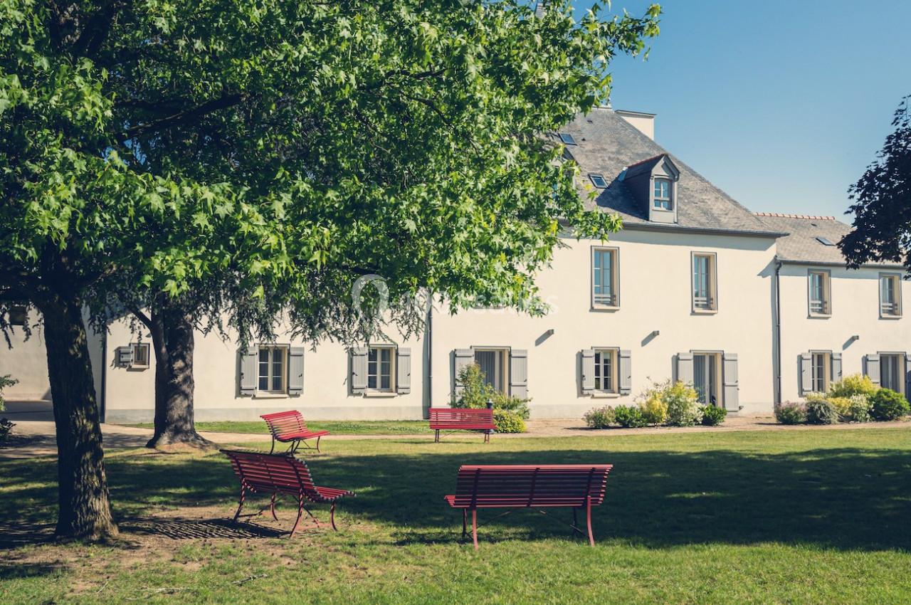 Bâtiment blanc à deux étages avec des volets gris, entouré d'arbres et de bancs rouges sur une pelouse verte.