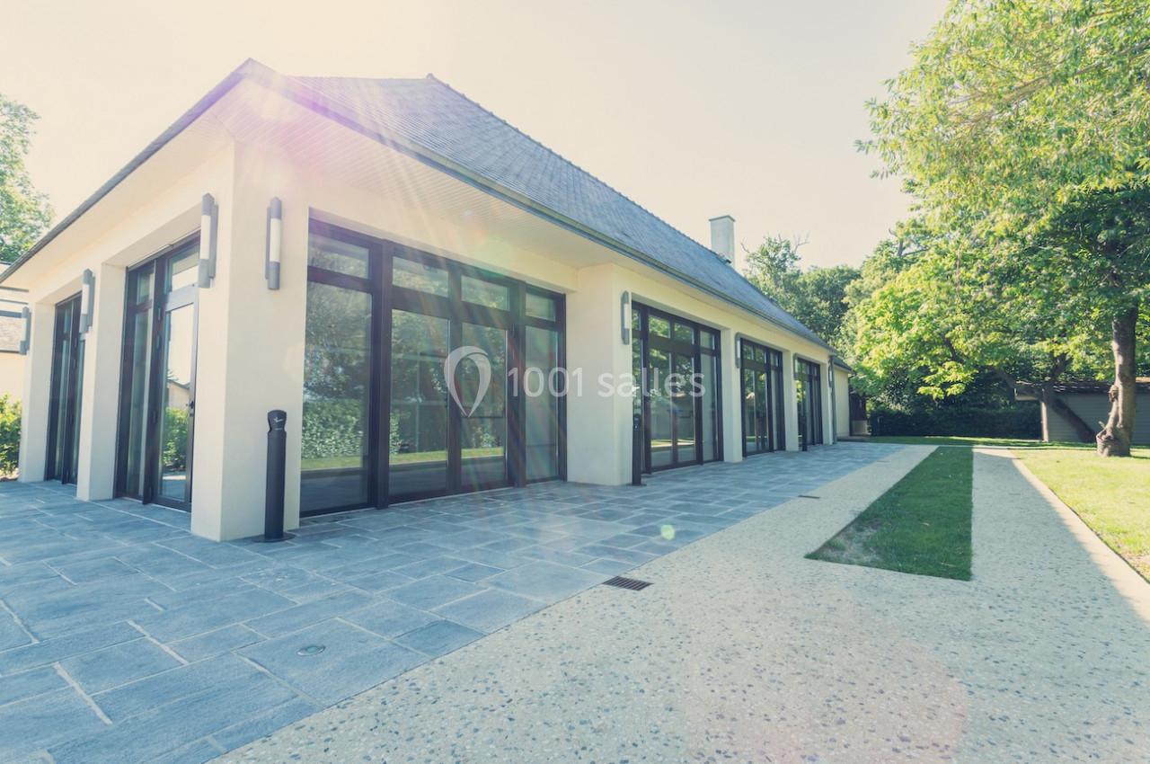 Maison moderne avec de grandes baies vitrées, entourée de végétation, vue depuis une allée pavée sous un ciel ensoleillé.