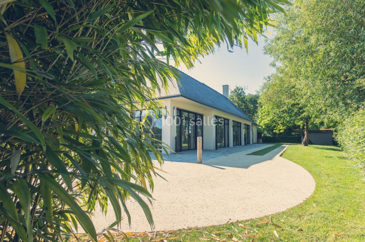 Façade d'une maison moderne avec de grandes baies vitrées, entourée de végétation et d'une allée en gravier.