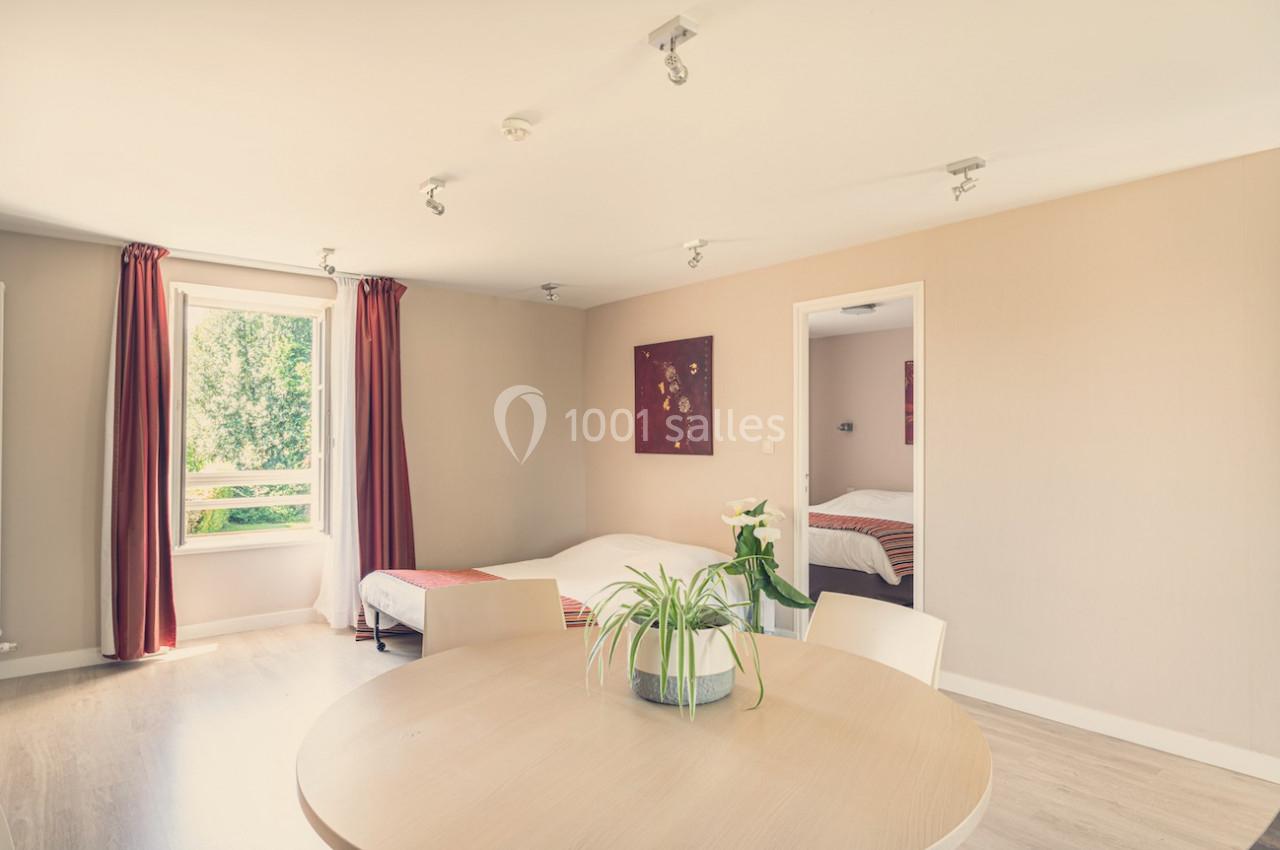 Chambre lumineuse avec deux lits, une table ronde, des rideaux bordeaux et une vue sur un espace vert.