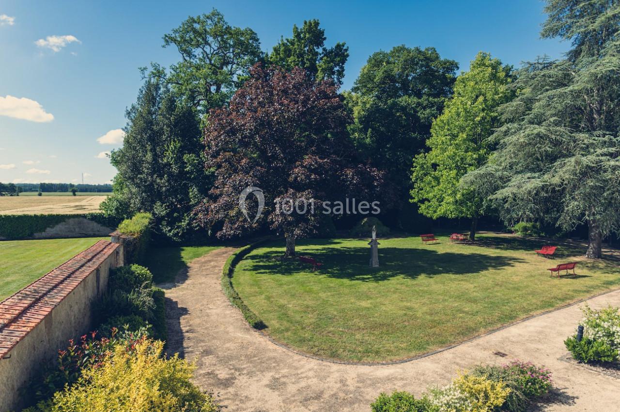 Jardin avec pelouse, arbres variés, bancs rouges et allées sinueuses sous un ciel bleu clair.