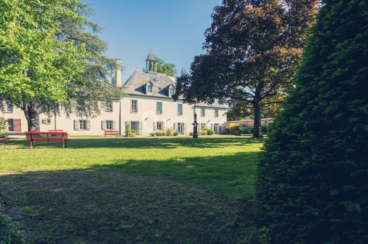 Vue d'un grand bâtiment ancien entouré d'un jardin arboré avec des bancs rouges sous un ciel dégagé.